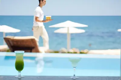 a man standing by a pool with a table with two glasses at Giannoulis - Grand Bay Beach Resort (Exclusive Adults Only) in Kolymvari