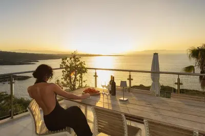 a woman sitting at a table on a balcony looking at the ocean at Villa Anton by Cura Villas Collection in Kalathas