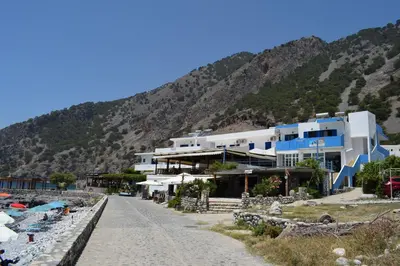 a building on a beach next to a mountain at Zorbas Studios in Agia Roumeli